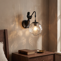 Vintage rustic black mermaid wall sconce with clear hammered glass orb glowing warm beside a walnut nightstand