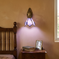 Vintage Tiffany purple stained glass petal wall sconce glowing warm beside a dark oak bed above an antique nightstand
