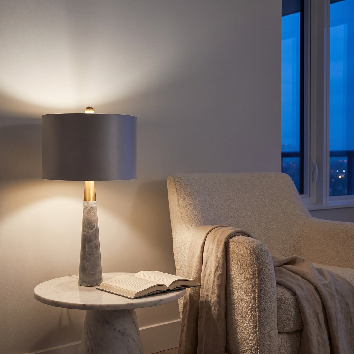 Pravelle stone and fabric table lamp lit at dusk beside a reading armchair