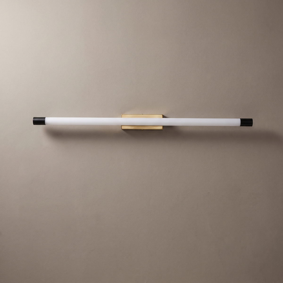 Thin aluminum PC tube LED wall sconce with gold center mount and black end caps on a greige plaster wall, lamp off