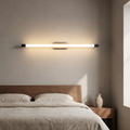 Modern minimalist thin tube LED wall sconce in black and gold glowing warm above a walnut bed headboard