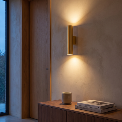 Gold cylindrical 2-light wall sconce lit at dusk in a minimalist living room above a walnut console, showing mounting height