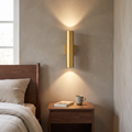 Modern minimalist gold aluminum cylindrical 2-light wall sconce glowing warm up and down beside a walnut bed and nightstand