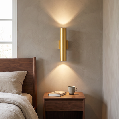 Modern minimalist gold aluminum cylindrical 2-light wall sconce glowing warm up and down beside a walnut bed and nightstand