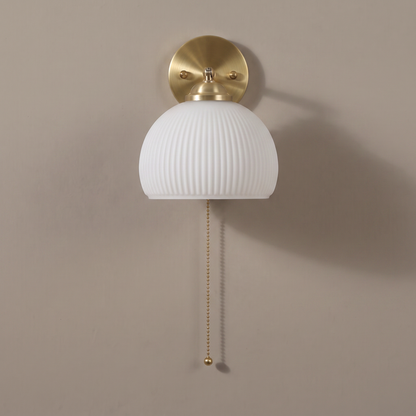 Contemporary ribbed white glass dome wall sconce with brass backplate and pull chain on a plain greige wall, lamp off