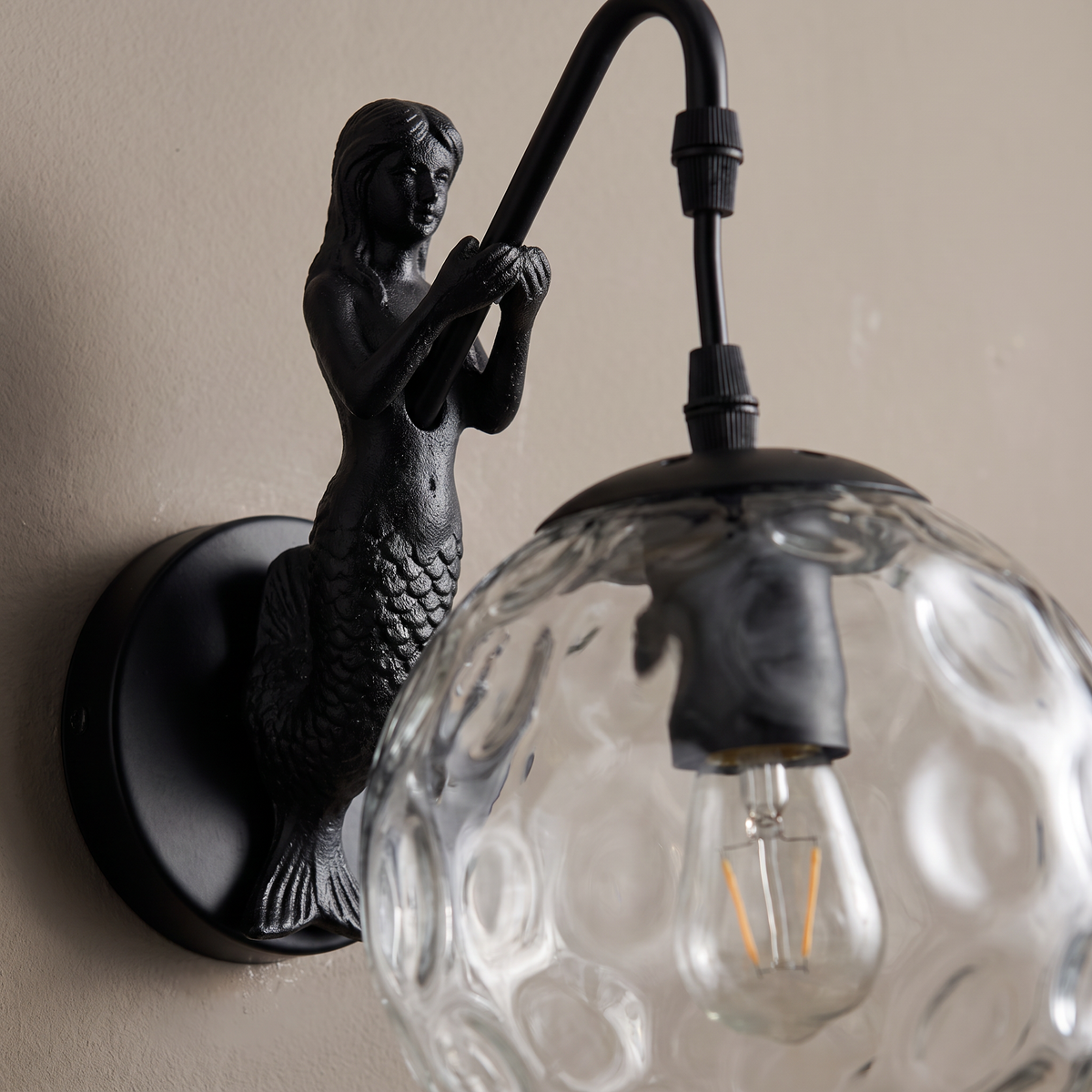 Close-up of the black mermaid figurine holding the arm above the rippled hammered glass orb shade of the wall sconce