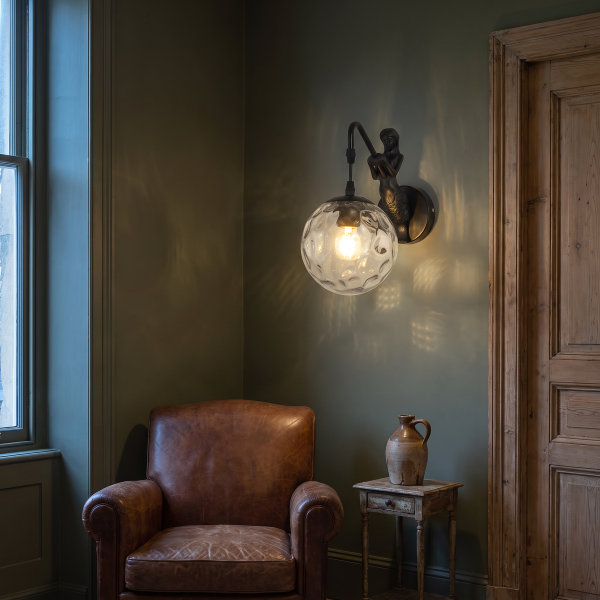 Mermaid orb wall sconce lit on an olive green wall beside a leather armchair in a vintage living room at dusk