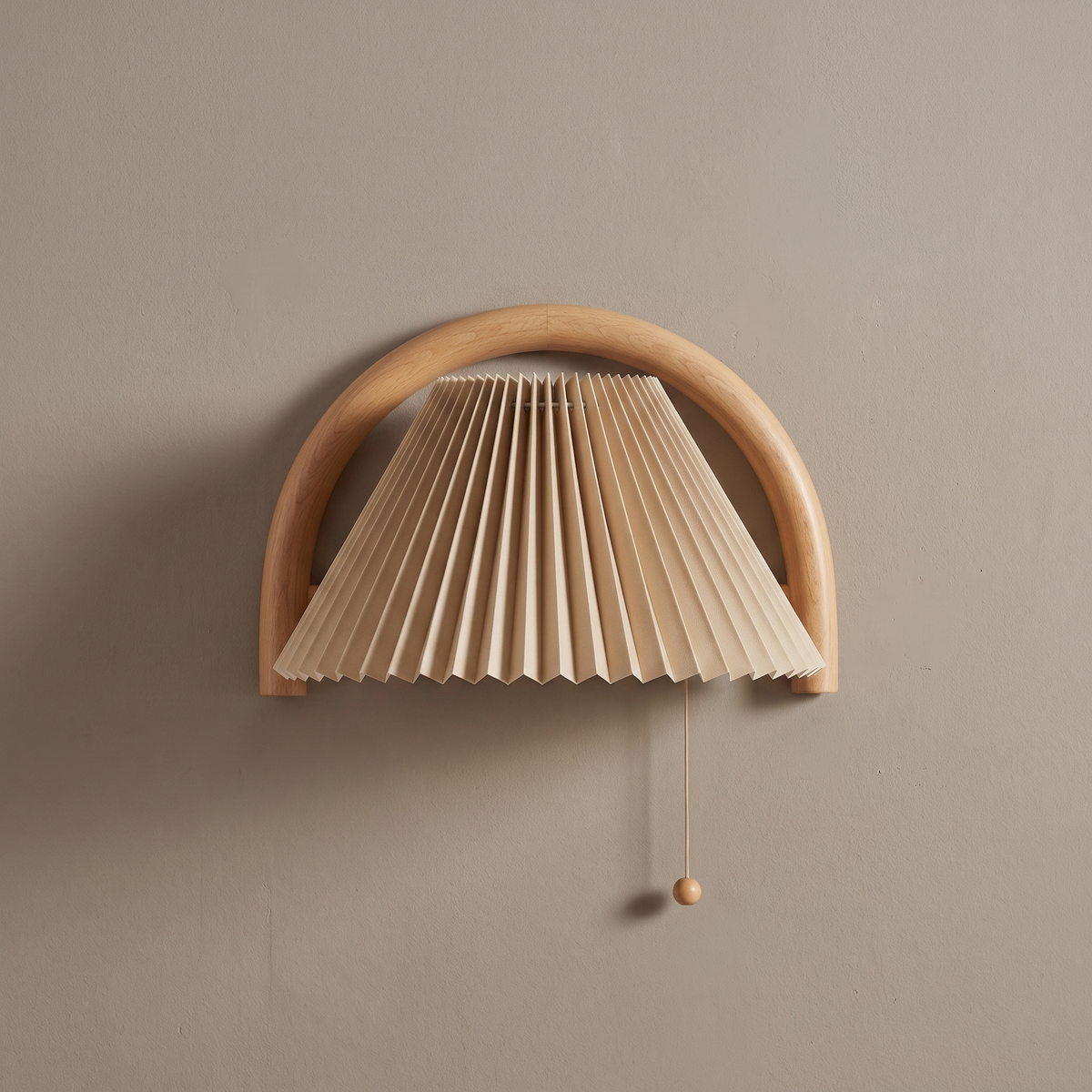 Solid wood semi-circle arch wall sconce with beige pleated shade and pull cord mounted on a greige plaster wall, lamp off