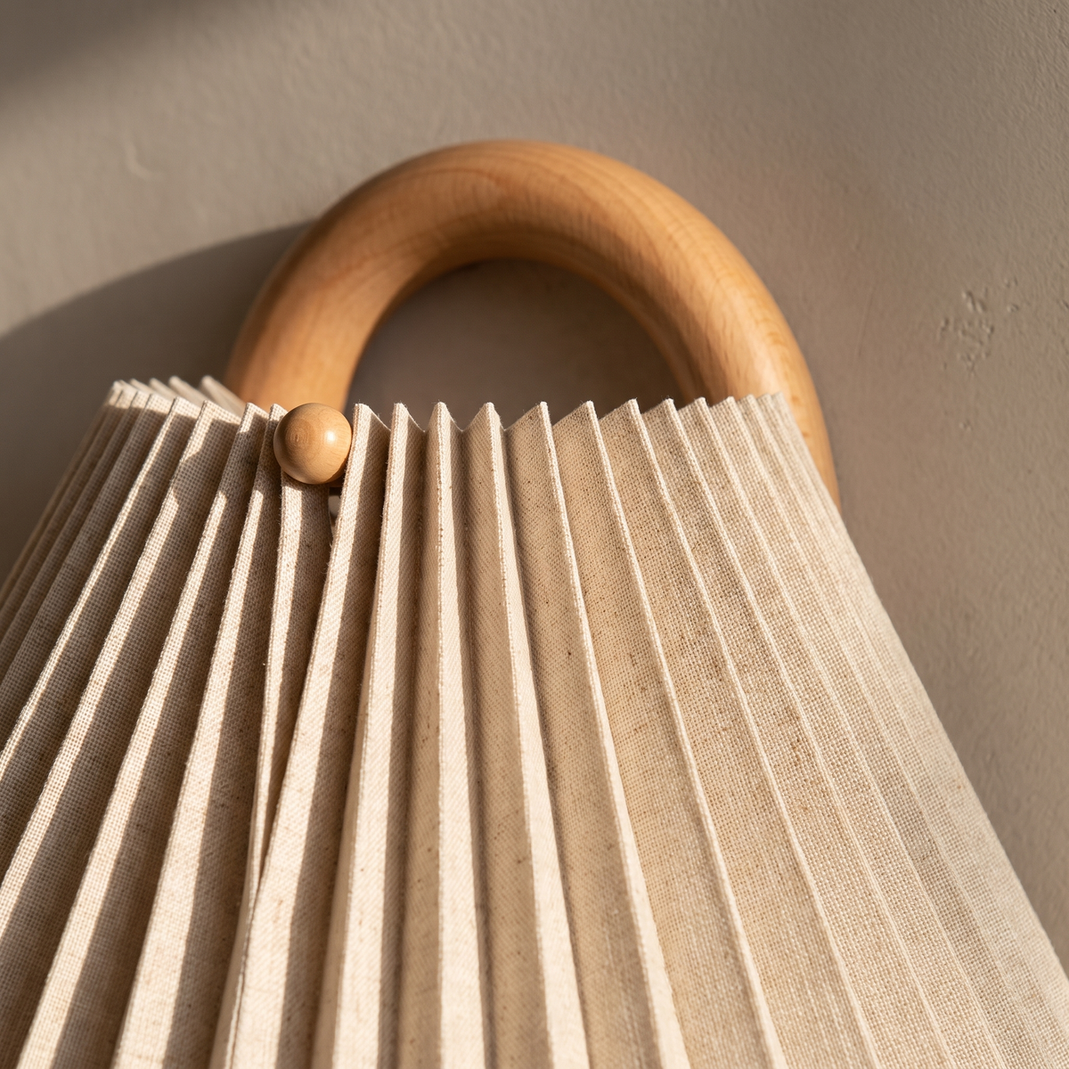 Close-up of the beige knife-pleated fabric shade meeting the natural beechwood arch of the wall sconce