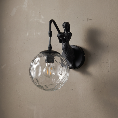 Black resin mermaid 1-light wall sconce with hammered clear glass orb mounted on a greige plaster wall, lamp off