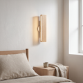 Nordic rotatable solid oak LED bar wall sconce glowing warm on a white plaster wall beside a bed with an oak nightstand