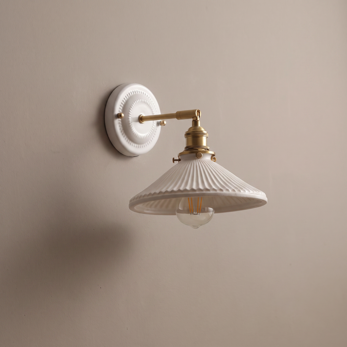 Vintage round ceramic backplate wall sconce with brass arm and white conical ceramic shade on a plain greige wall, lamp off