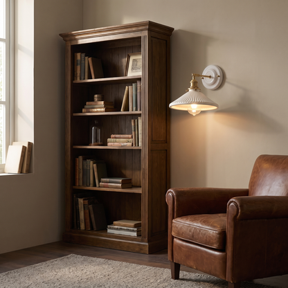 Traditional vintage white ceramic and brass 1-light wall sconce glowing warm above a leather armchair beside an oak bookcase
