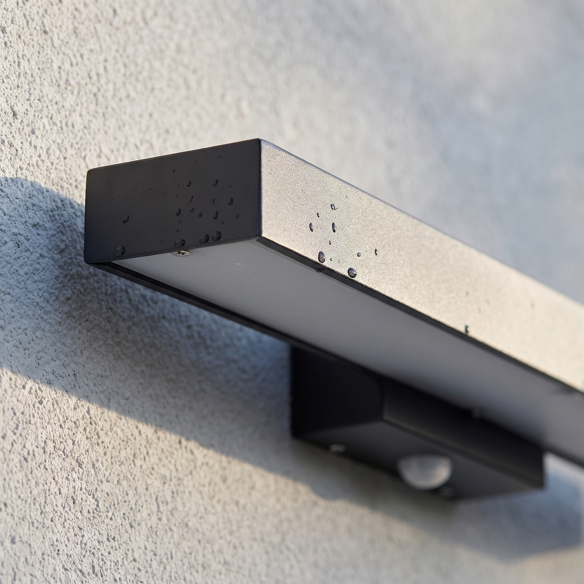 Close-up of the matte black stainless steel housing, frosted diffuser and motion sensor of the outdoor wall sconce with rain droplets