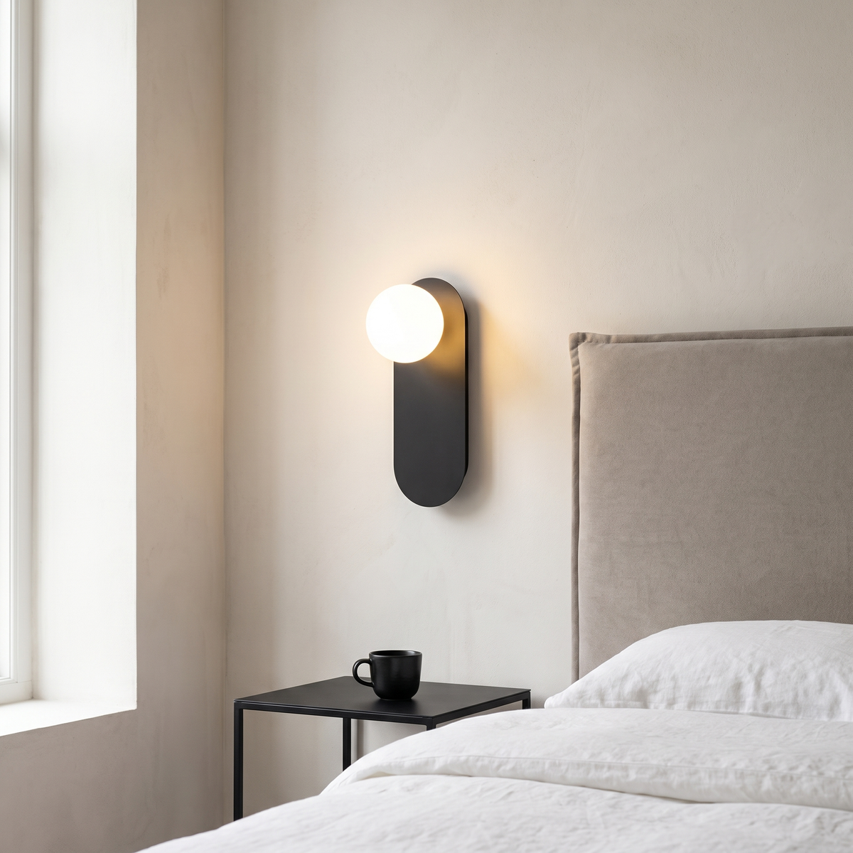 Nordic minimalist black pill-shaped glass globe wall sconce glowing warm beside a greige headboard