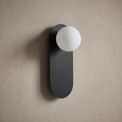 Matte black capsule backplate wall sconce with white opal glass globe on a plain greige wall, light off