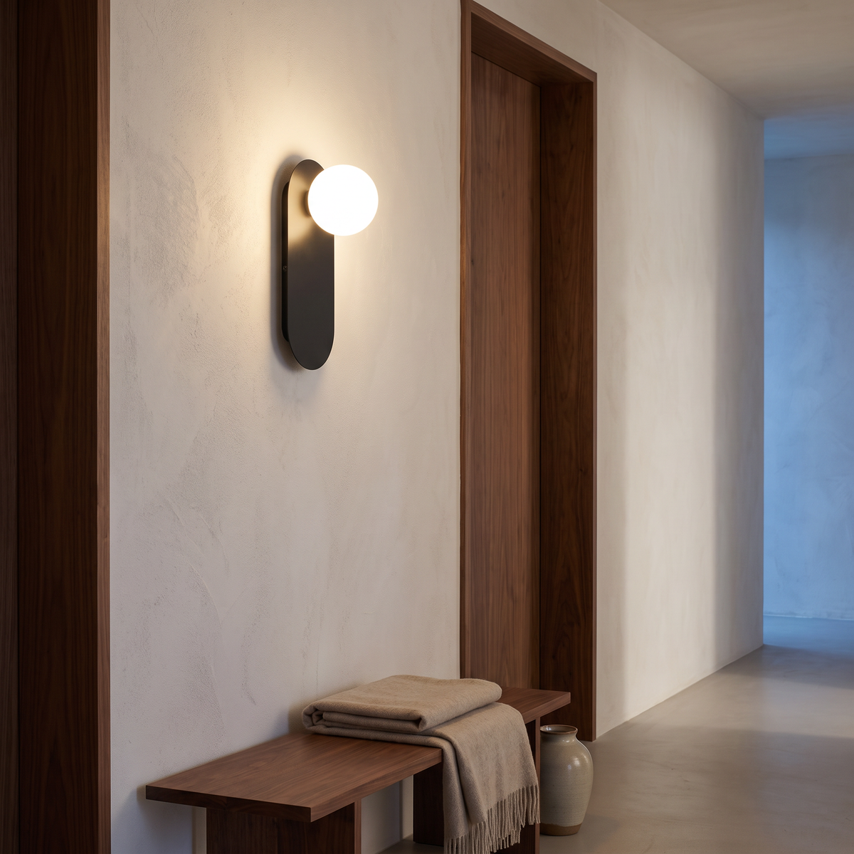 Black minimalist glass globe wall sconce lit in a warm hallway beside a walnut door frame and bench