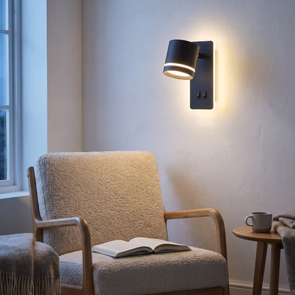 Black rotatable spotlight LED wall sconce lit above a reading armchair in a Nordic living room at dusk