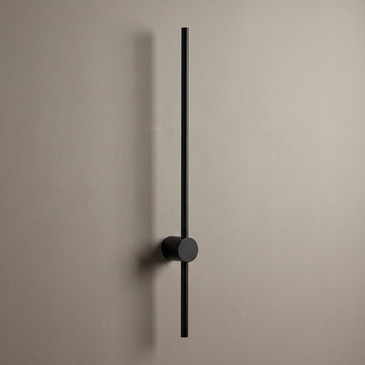 Thin black aluminum vertical strip LED wall sconce with round wall mount on a greige plaster wall, lamp off
