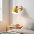 Macaron yellow iron and wood wall sconce with pull cord switch glowing warm beside an oak bed