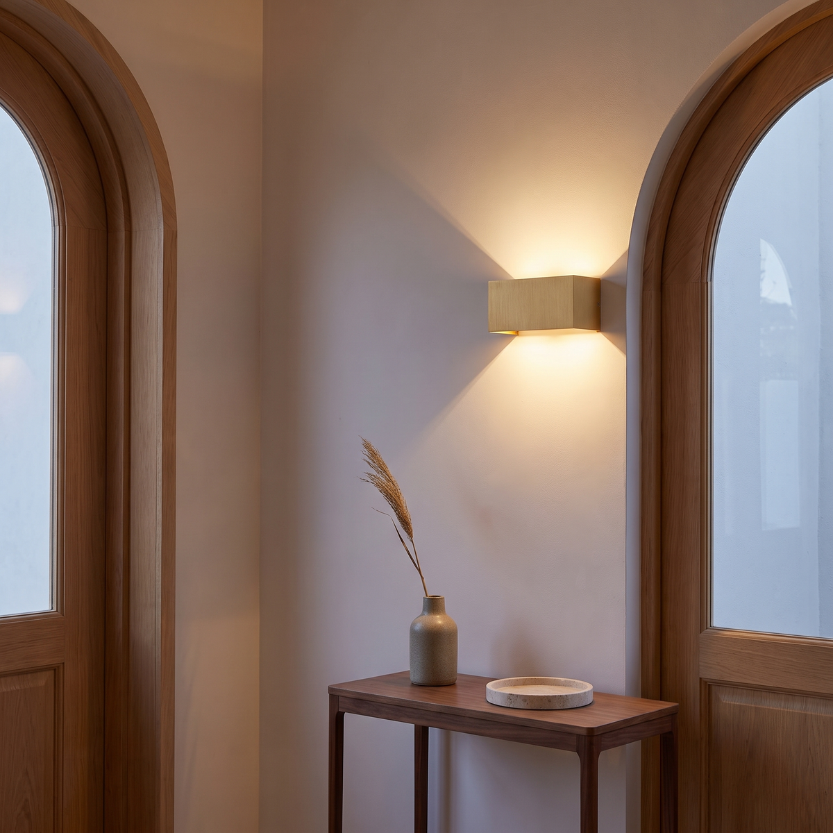 Gold aluminum minimalist LED wall sconce lit in an entryway beside an arched oak doorway above a walnut console, showing mounting height