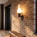 Retro kerosene style antique bronze wall sconce with frosted glass chimney glowing warm on a reclaimed brick hallway wall