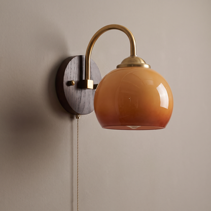Mid-century caramel glass dome wall sconce with round ash wood backplate, brass gooseneck arm and pull chain on a plain greige wall, light off