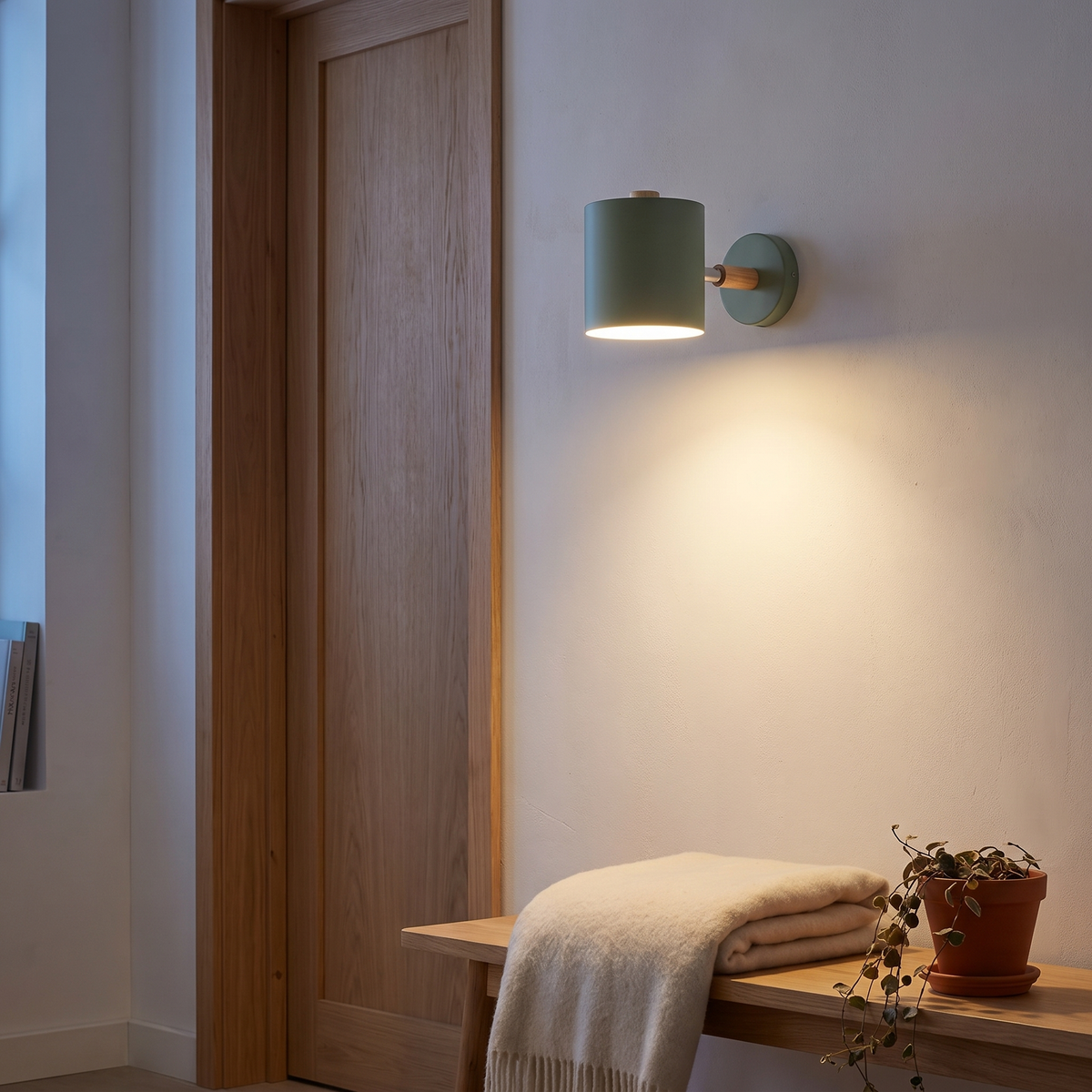 Nordic macaron sage green cylinder wall sconce lit beside an oak door frame above a bench in a Scandinavian reading nook, showing scale