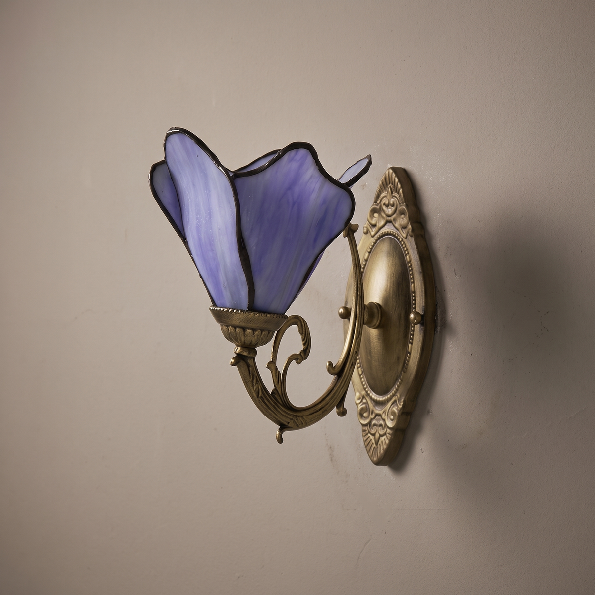 Tiffany style purple stained glass 1-light wall sconce with antique bronze scroll arm, lamp off on a plaster wall