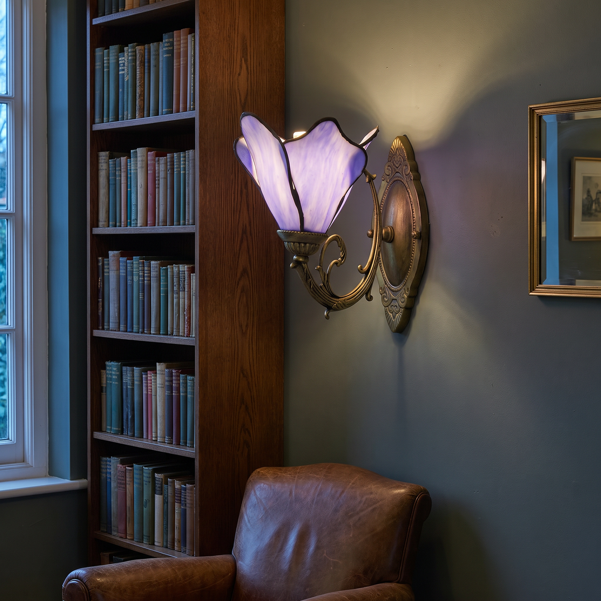 Vintage Tiffany petal stained glass wall lamp lit in a study reading corner beside a bookcase and leather armchair