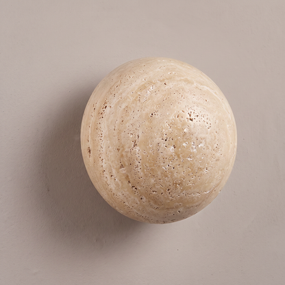 Round yellow travertine stone disc wall sconce on a plain greige plaster wall, lamp off, showing natural pitted texture