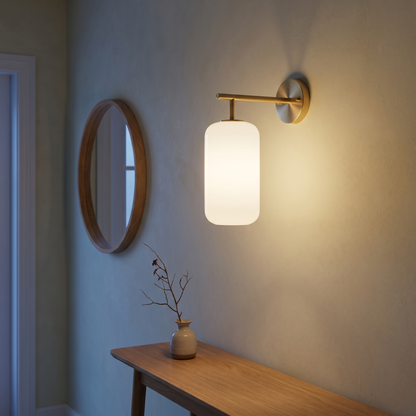 Brass and lucite cylinder wall sconce lit at dusk in a Japandi entry hallway beside an oak console and round mirror, showing 15 cm arm projection and mounting height