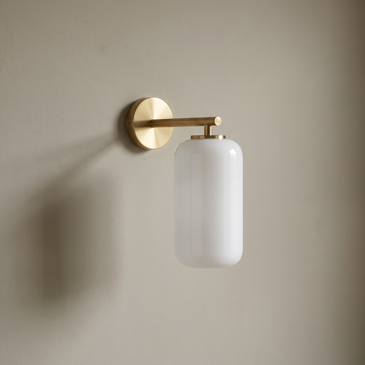 Minimalist brushed brass straight-arm wall sconce with white translucent lucite cylinder shade mounted on a plain greige plaster wall, lamp off