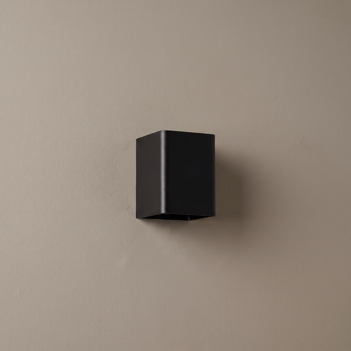 Small matte black square aluminum LED wall sconce mounted on a plain greige plaster wall, lamp off, showing compact cube form