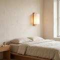 Modern minimalist solid wood and frosted glass column wall sconce glowing warm above an oak nightstand in a Japandi bedroom